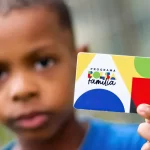 Imagem colorida mostra criança segurando cartão do Bolsa Família