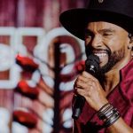 Alexandre Pires sorri para a câmera, cantando no palco, usando chapéu e camisa vermelha.