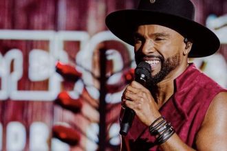 Alexandre Pires sorri para a câmera, cantando no palco, usando chapéu e camisa vermelha.