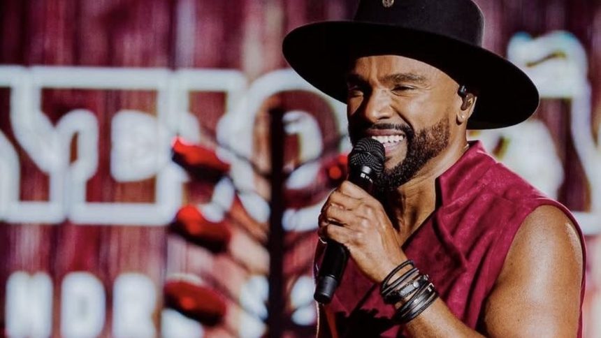 Alexandre Pires sorri para a câmera, cantando no palco, usando chapéu e camisa vermelha.