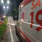 Ambulância do SAMU 192 estacionada em uma estrada, ao lado de um corpo coberto por um lençol branco.