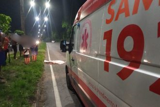 Ambulância do SAMU 192 estacionada em uma estrada, ao lado de um corpo coberto por um lençol branco.