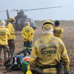 Brigadistas do ICMBio, com uniformes amarelos, observam um helicóptero durante operação de combate a incêndios florestais.