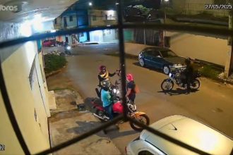 Câmera de segurança mostra dois indivíduos em uma moto sendo abordados por três assaltantes em uma rua à noite.