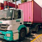 Caminhão branco com a logomarca 'Super Trans' e a inscrição 'Axor' estacionado em um pátio de containers, com um container rosa com a inscrição 'ONE' atrás dele.