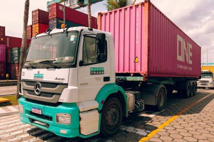 Caminhão branco com a logomarca 'Super Trans' e a inscrição 'Axor' estacionado em um pátio de containers, com um container rosa com a inscrição 'ONE' atrás dele.