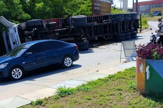 Carreta tombada na estrada com um carro azul parado próximo. O acidente ocorreu na Estrada da Colônia Antônio Aleixo, em frente ao Centro do Alumínio.