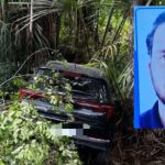 Carro acidentado em uma ribanceira com árvores, próximo a um cartaz com a foto de um homem.