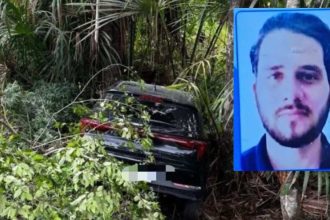 Carro acidentado em uma ribanceira com árvores, próximo a um cartaz com a foto de um homem.