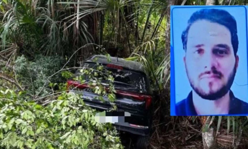 Carro acidentado em uma ribanceira com árvores, próximo a um cartaz com a foto de um homem.