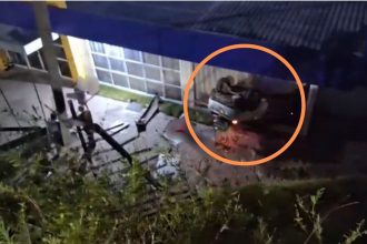 Carro branco acidentado no chão com a frente amassada após cair de um barranco, na área externa de uma escola.