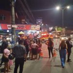 Cena de um acidente fatal em uma rua noturna, com um homem morto no chão, pessoas reunidas, um carro da polícia e uma ambulância.