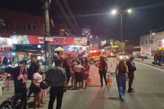Cena de um acidente fatal em uma rua noturna, com um homem morto no chão, pessoas reunidas, um carro da polícia e uma ambulância.