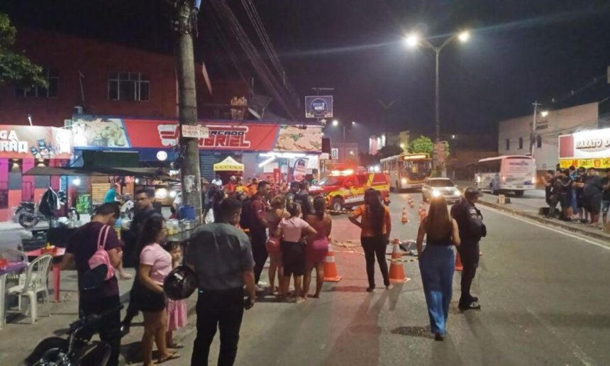 Cena de um acidente fatal em uma rua noturna, com um homem morto no chão, pessoas reunidas, um carro da polícia e uma ambulância.