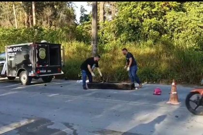 Corpos de policiais forenses carregando um caixão preto próximo a um veículo do IML, com uma motocicleta caída e cones laranja no chão.