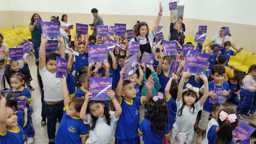 Crianças segurando livros infantis em uma sala de aula. A imagem mostra um grupo de crianças pequenas em uma sala de aula, todas segurando livros infantis e sorrindo para a câmera.