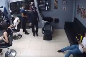 Dois homens em uma barbearia. Um dos homens está sendo preso por um policial. O outro homem está sentado em um sofá e observa a cena.