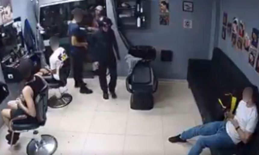 Dois homens em uma barbearia. Um dos homens está sendo preso por um policial. O outro homem está sentado em um sofá e observa a cena.
