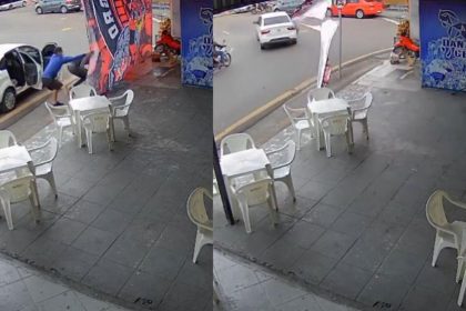 Dois homens retiram uma bandeira do Flamengo de um poste em frente a um bar, com um carro branco estacionado próximo.