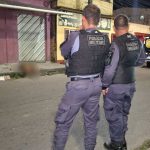 Dois policiais militares em frente a um corpo coberto por um lençol branco, no chão de uma rua, durante um suposto crime.