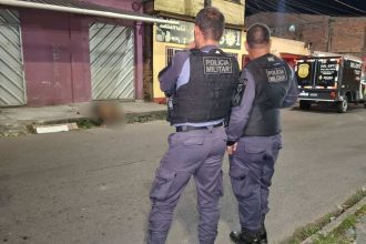 Dois policiais militares em frente a um corpo coberto por um lençol branco, no chão de uma rua, durante um suposto crime.