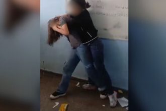 Duas meninas em uma sala de aula, uma segurando a outra pelo braço enquanto a outra está com a cabeça baixa.