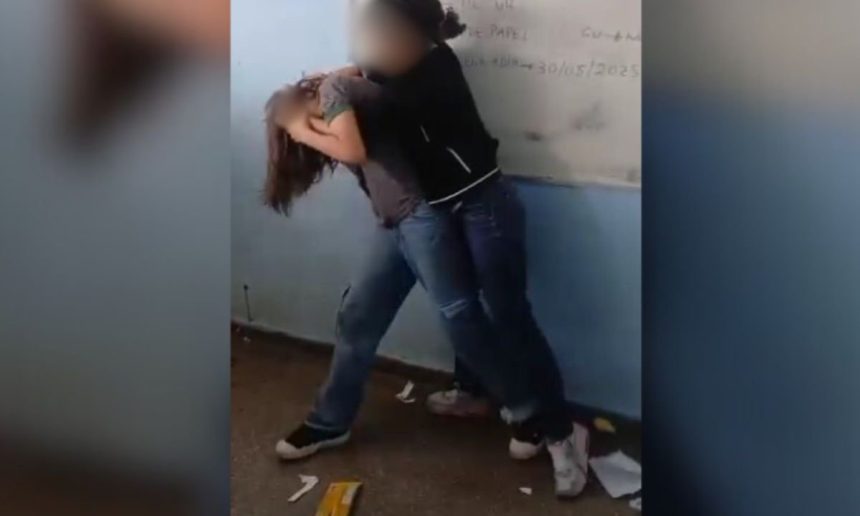 Duas meninas em uma sala de aula, uma segurando a outra pelo braço enquanto a outra está com a cabeça baixa.