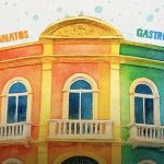 Fachada colorida de um prédio com detalhes em arco, janelas e varandas. A fachada tem o arco decorado com as palavras "Artesanatos", "Gastronomia", "Bebidas" e "Receitas", em destaque.