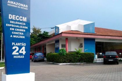 Fachada da Delegacia Especializada em Crimes Contra a Mulher (DECCM) em Manaus, Amazonas, com a placa de plantão 24 horas.