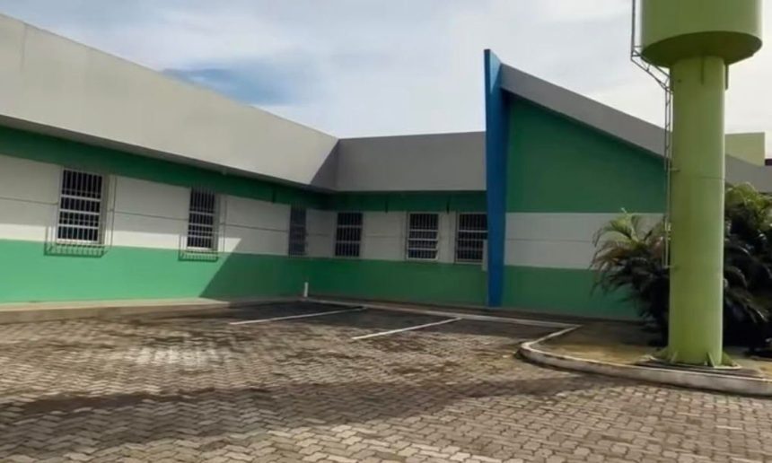 Fachada de uma clínica com paredes verdes e brancas, um estacionamento e um tanque de água alto.