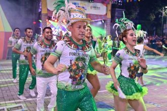 Grupo de dançarinos em trajes coloridos com plumas e fantasias, participando de um evento de festival folclórico, em Manaus.