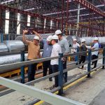 Grupo de pessoas em uma fábrica, possivelmente durante uma visita ou inspeção, observando rolos de metal.