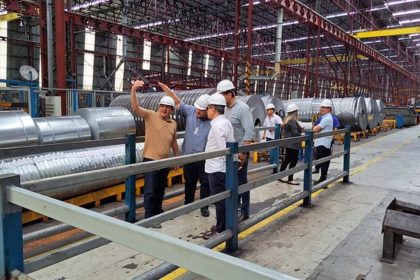 Grupo de pessoas em uma fábrica, possivelmente durante uma visita ou inspeção, observando rolos de metal.