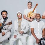 Grupo musical de seis homens, vestidos de branco, tocando instrumentos musicais. Um deles segura um violão, outro um banjo e outro um pandeiro.