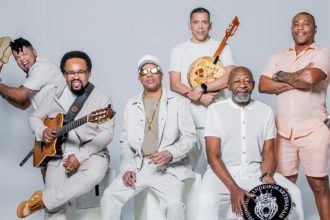 Grupo musical de seis homens, vestidos de branco, tocando instrumentos musicais. Um deles segura um violão, outro um banjo e outro um pandeiro.