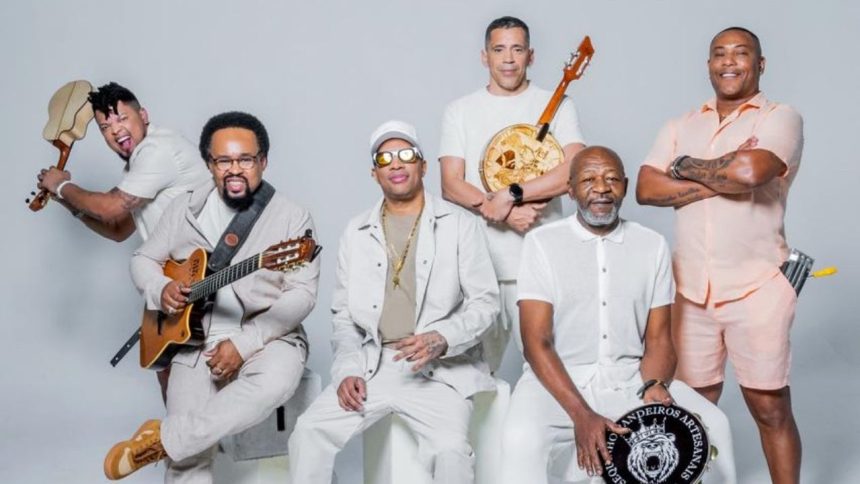 Grupo musical de seis homens, vestidos de branco, tocando instrumentos musicais. Um deles segura um violão, outro um banjo e outro um pandeiro.