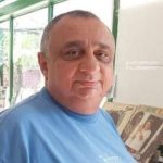 Homem de meia idade com cabelos grisalhos e barba, vestindo camisa azul, em um ambiente interno com janelas e um fundo desfocado de outros objetos.