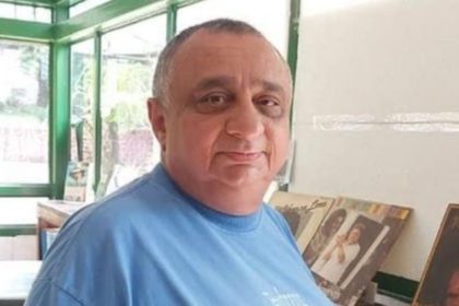 Homem de meia idade com cabelos grisalhos e barba, vestindo camisa azul, em um ambiente interno com janelas e um fundo desfocado de outros objetos.