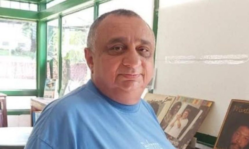 Homem de meia idade com cabelos grisalhos e barba, vestindo camisa azul, em um ambiente interno com janelas e um fundo desfocado de outros objetos.