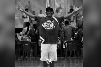 Homem sorridente vestindo camiseta do Boi-Bumbá Garantido com os braços abertos, em frente a um público em um evento.