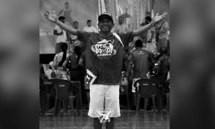 Homem sorridente vestindo camiseta do Boi-Bumbá Garantido com os braços abertos, em frente a um público em um evento.