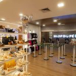 Interior de uma loja de calçados com prateleiras cheias de sapatos, um balcão com a marca 'Shop do Pé' e algumas pessoas.