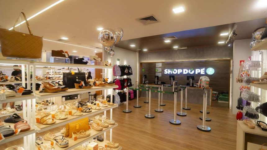 Interior de uma loja de calçados com prateleiras cheias de sapatos, um balcão com a marca 'Shop do Pé' e algumas pessoas.
