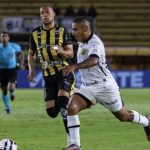 Jogadores de futebol em campo, um em uniforme branco e outro em uniforme listrado amarelo e preto.