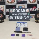 Mesa com objetos apreendidos de suspeitos de roubo, incluindo placa de carro, celular, cartão, faca e símbolo policial.