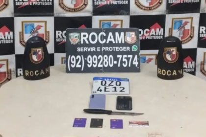 Mesa com objetos apreendidos de suspeitos de roubo, incluindo placa de carro, celular, cartão, faca e símbolo policial.