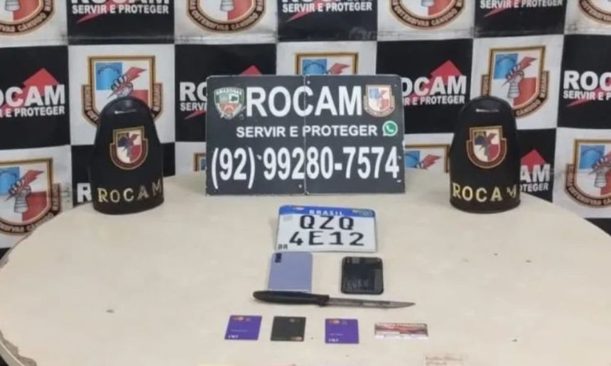 Mesa com objetos apreendidos de suspeitos de roubo, incluindo placa de carro, celular, cartão, faca e símbolo policial.