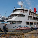 Navio de passageiros atracado no porto de Manaus, com pessoas desembarcando e a escrita "Parintins" visível na lateral, sugerindo viagem para o festival.