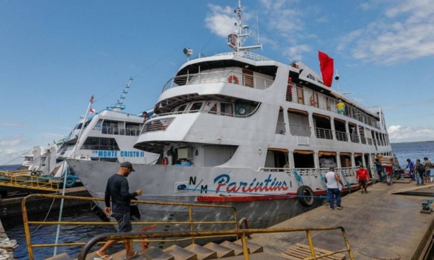 Navio de passageiros atracado no porto de Manaus, com pessoas desembarcando e a escrita "Parintins" visível na lateral, sugerindo viagem para o festival.
