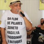 Paulo Onça, sambista, em um evento com um microfone na mão, usando uma camiseta branca com a frase "Da Flor de Pilares Guineto Magalha Pagodinho & Batuques".